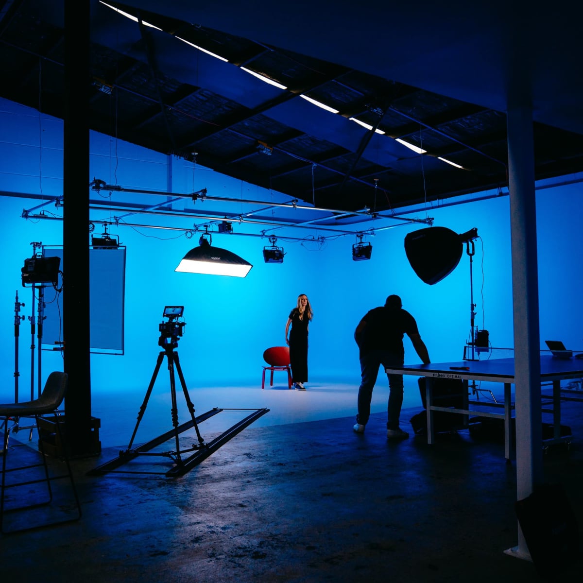 A person stands in front of a blue backdrop in a well-lit studio with camera equipment and lighting setups.