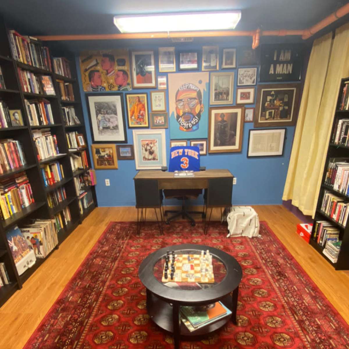 The room features shelves filled with books and artwork on the walls, a table with a chessboard in the center, and a chair positioned at a desk against a blue backdrop.