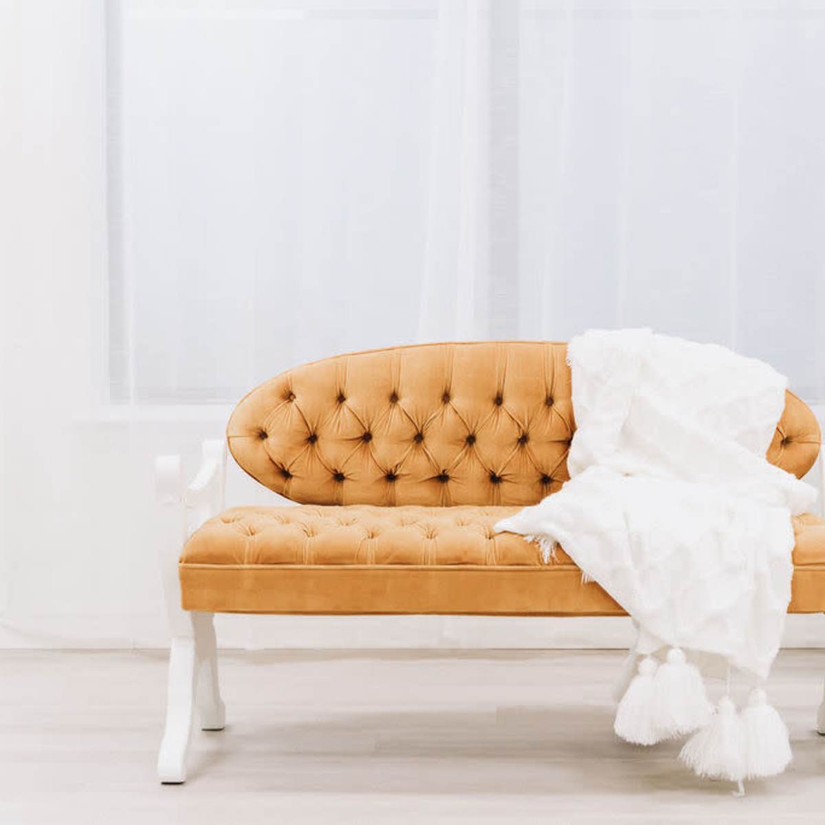 A tufted mustard-colored bench with a white blanket draped over it is positioned next to a decorative screen in a bright room.