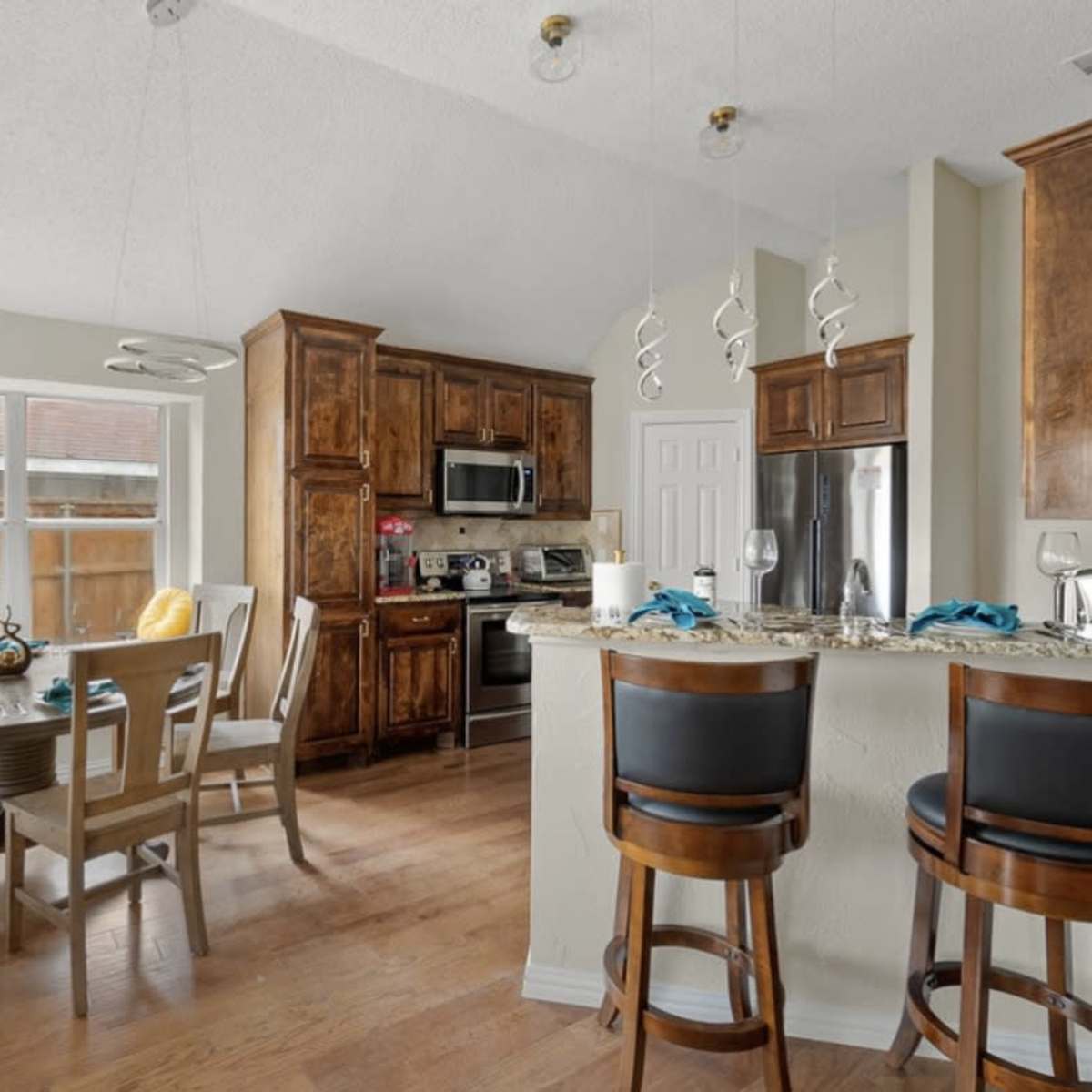 A kitchen features wooden cabinets, stainless steel appliances, and a dining area with a round table and bar stools.