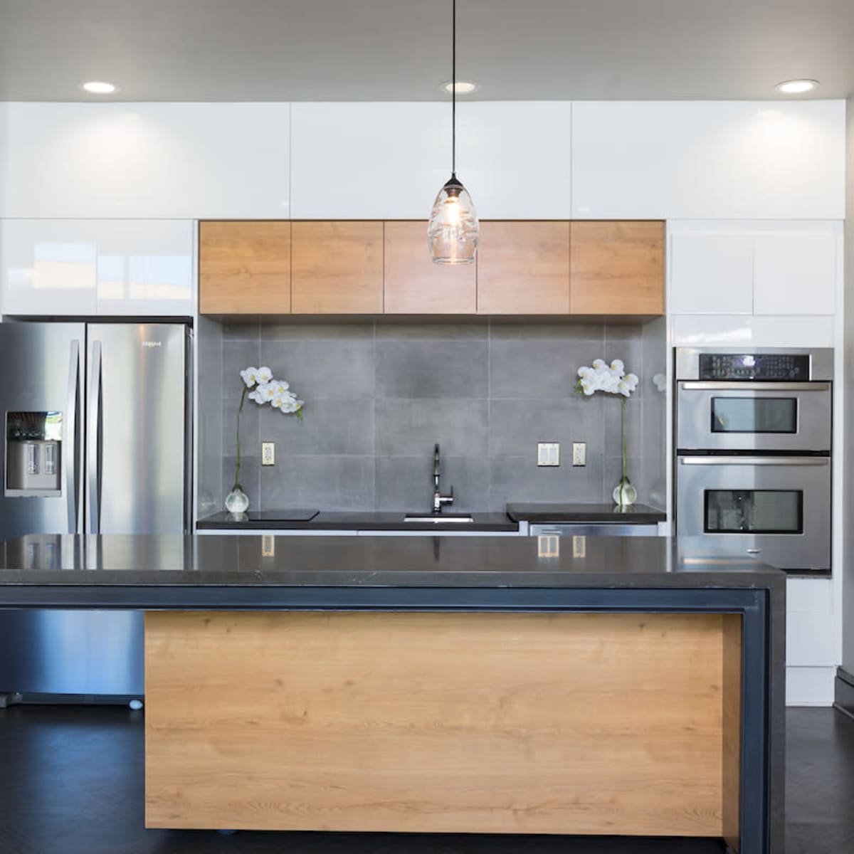 The kitchen features a central wooden island, stainless steel appliances, and a gray tiled backsplash with modern light fixtures above.