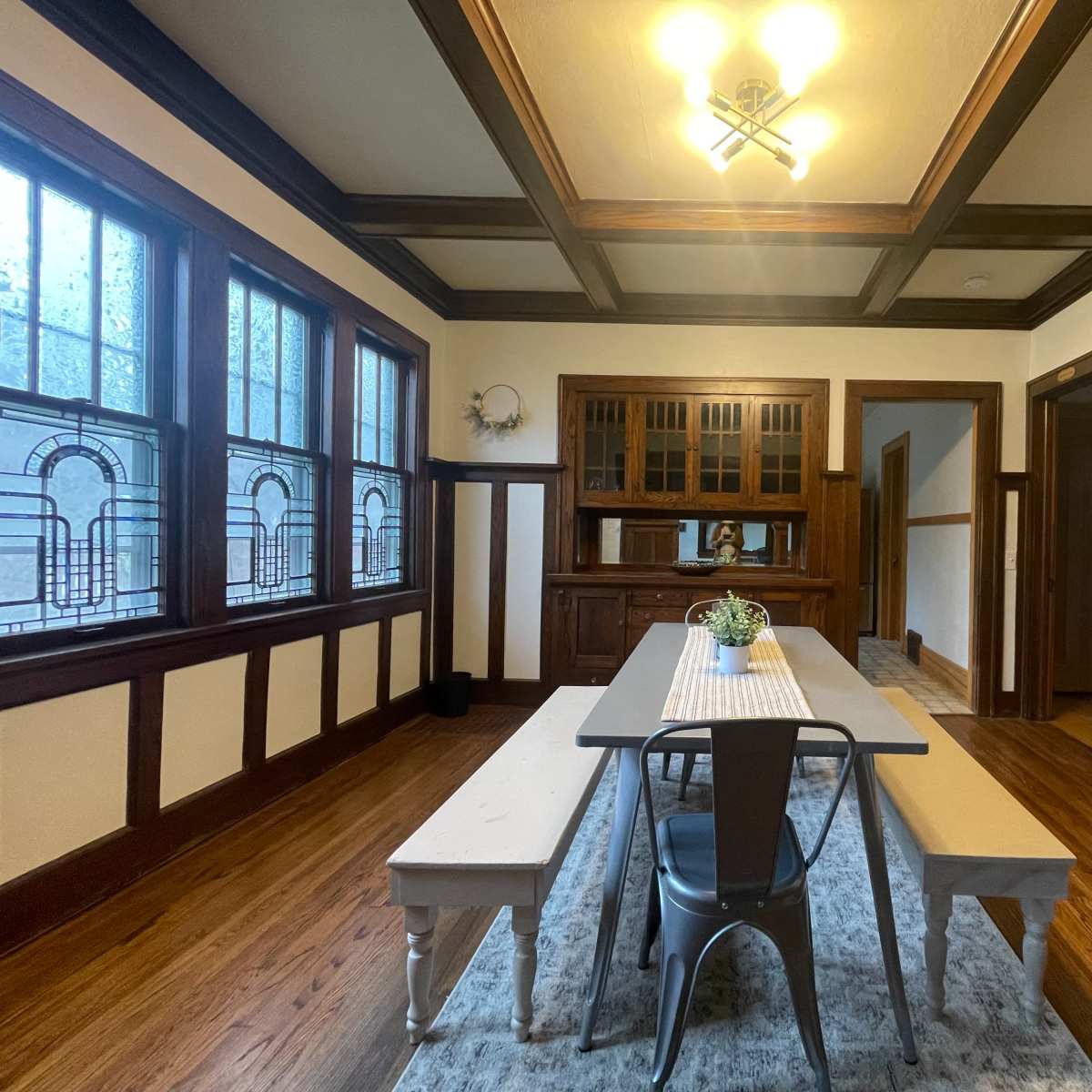 A dining room with a long table and benches situated under a coffered ceiling, surrounded by dark wooden paneling and large windows.