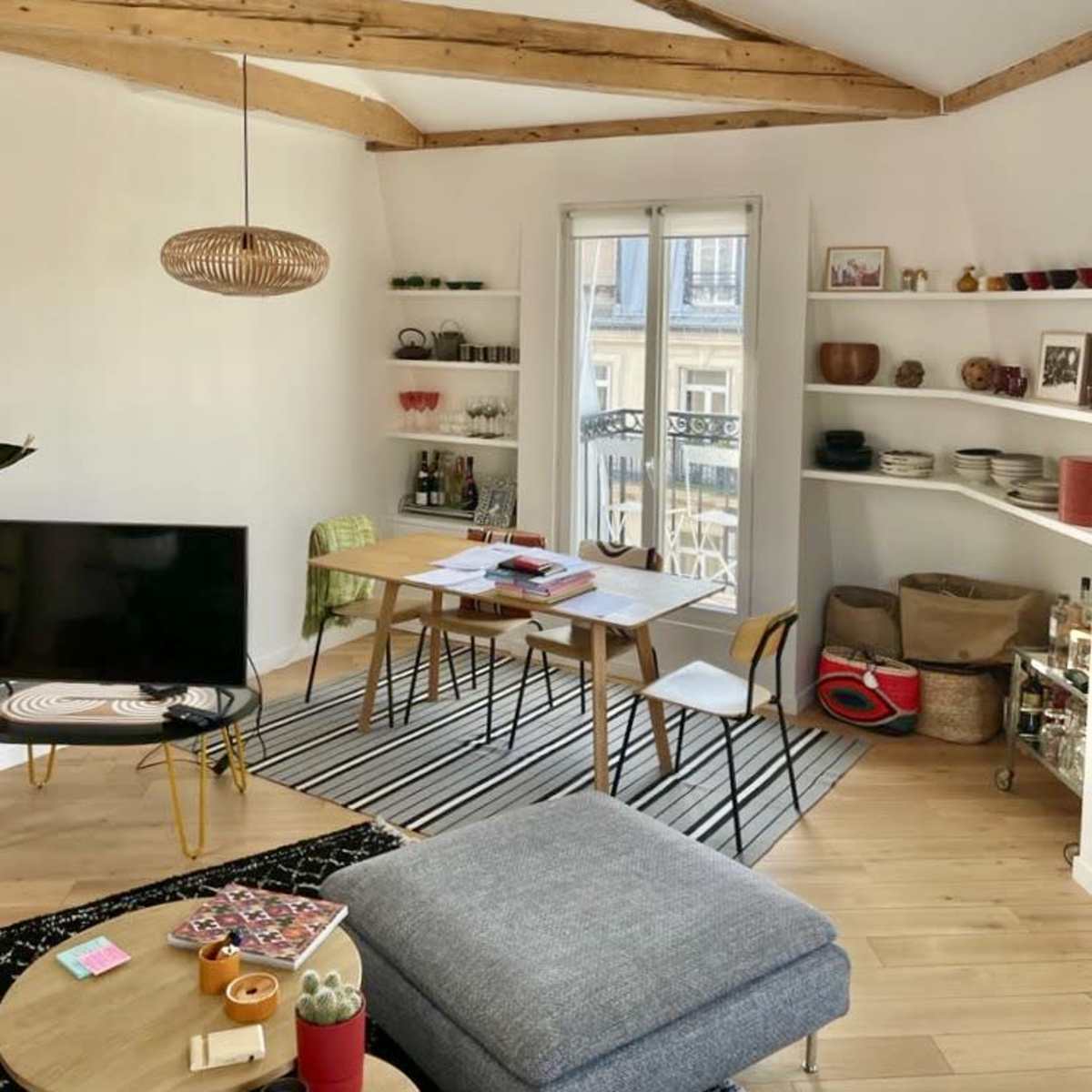 A bright living room features a television, a dining table with chairs, and shelves displaying various items, all under a vaulted ceiling with exposed beams.