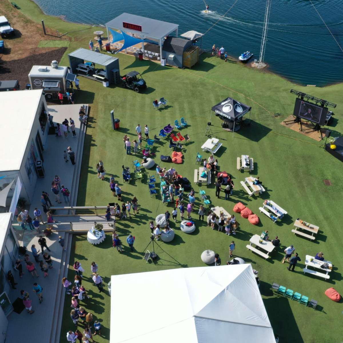 An aerial view shows a gathering of people on green lawn near a body of water, with tents, picnic tables, and various setups for activities.