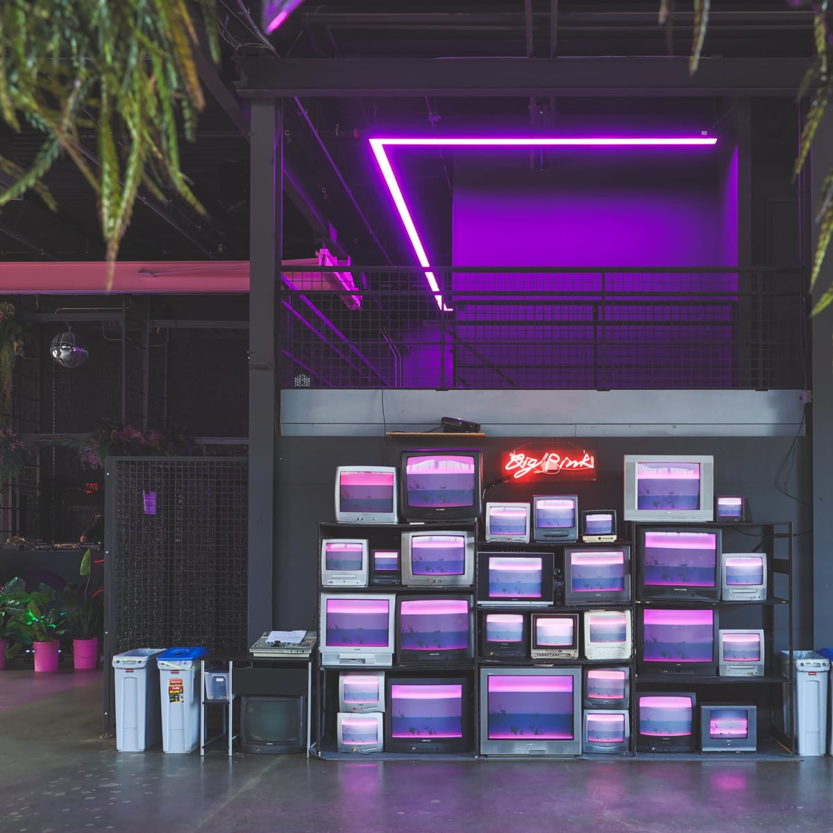 A wall of retro computer monitors is arranged in a grid, illuminated by pink neon lights in a modern indoor space.