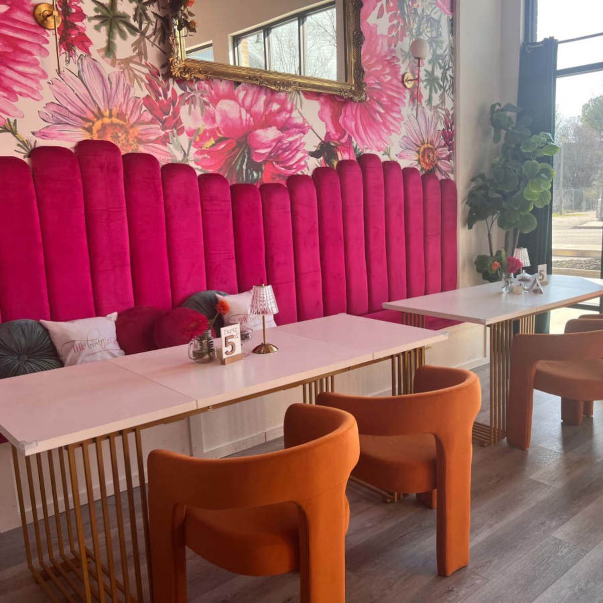 A brightly decorated café with pink floral wallpaper, two tables with white tops and gold legs, and orange velvet chairs.