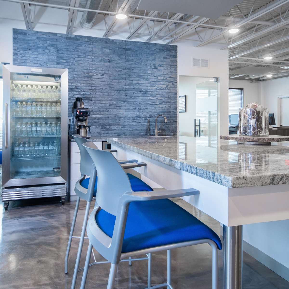 A modern office break area featuring a granite-topped counter with blue chairs, a refrigerator, and a coffee machine against a textured blue wall.