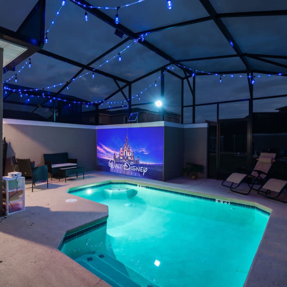 A backyard pool area with a glowing blue pool, a table and chairs, and a large screen displaying a Disney theme set against a nighttime sky.
