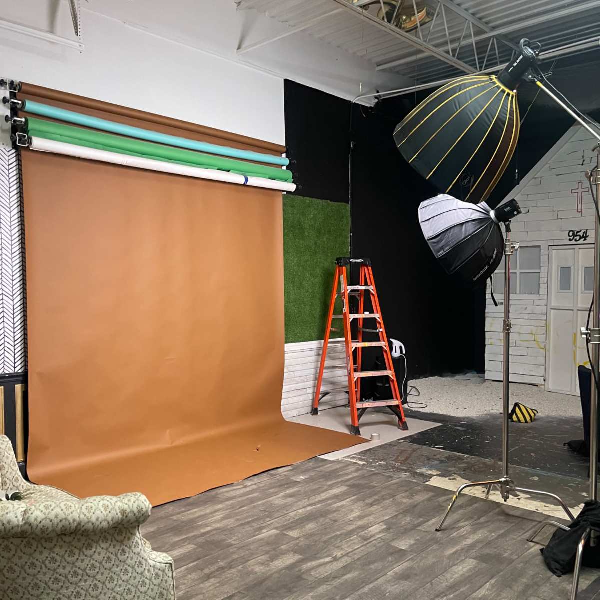 A photography studio with a brown backdrop, green and light-colored backdrop rolls, lighting equipment, an orange ladder, and a patterned armchair.