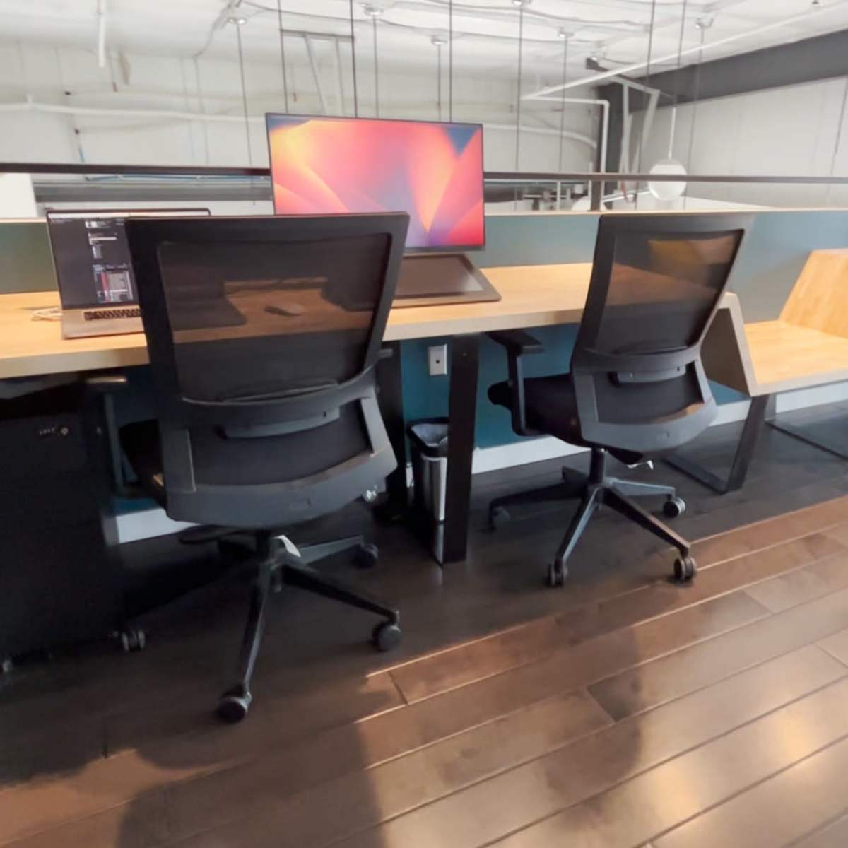 A row of modern office desks equipped with computer monitors and ergonomic chairs is set against a teal wall in a spacious workspace.
