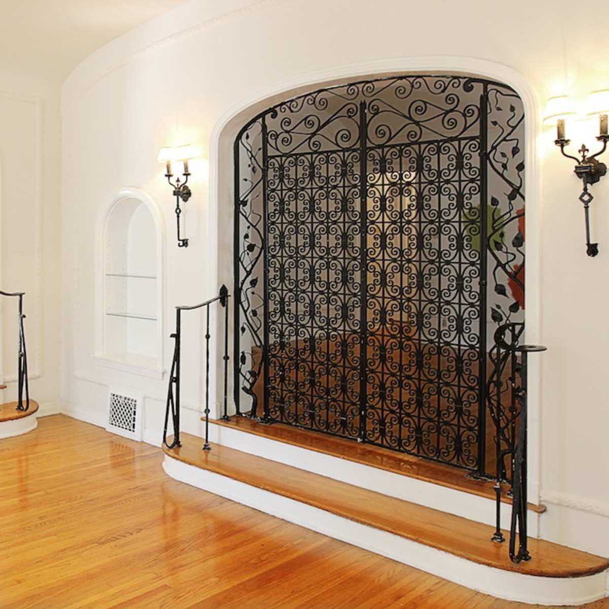 A spacious room featuring two wooden staircases leading to an ornate wrought iron gate, with warm wall sconces illuminating the space.