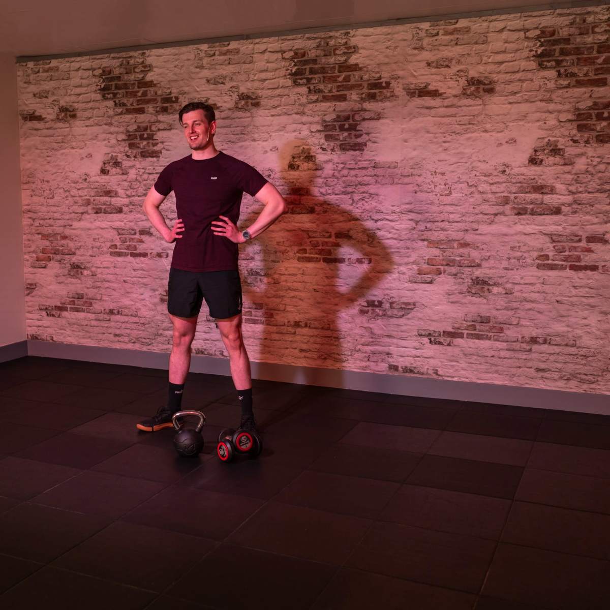 A man stands with his hands on his hips in a well-lit room, next to a kettlebell and a set of dumbbells on the floor.