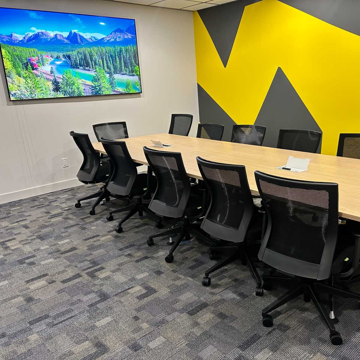 A large conference room features a long wooden table surrounded by black mesh chairs, with a large television displaying a scenic landscape on the wall.