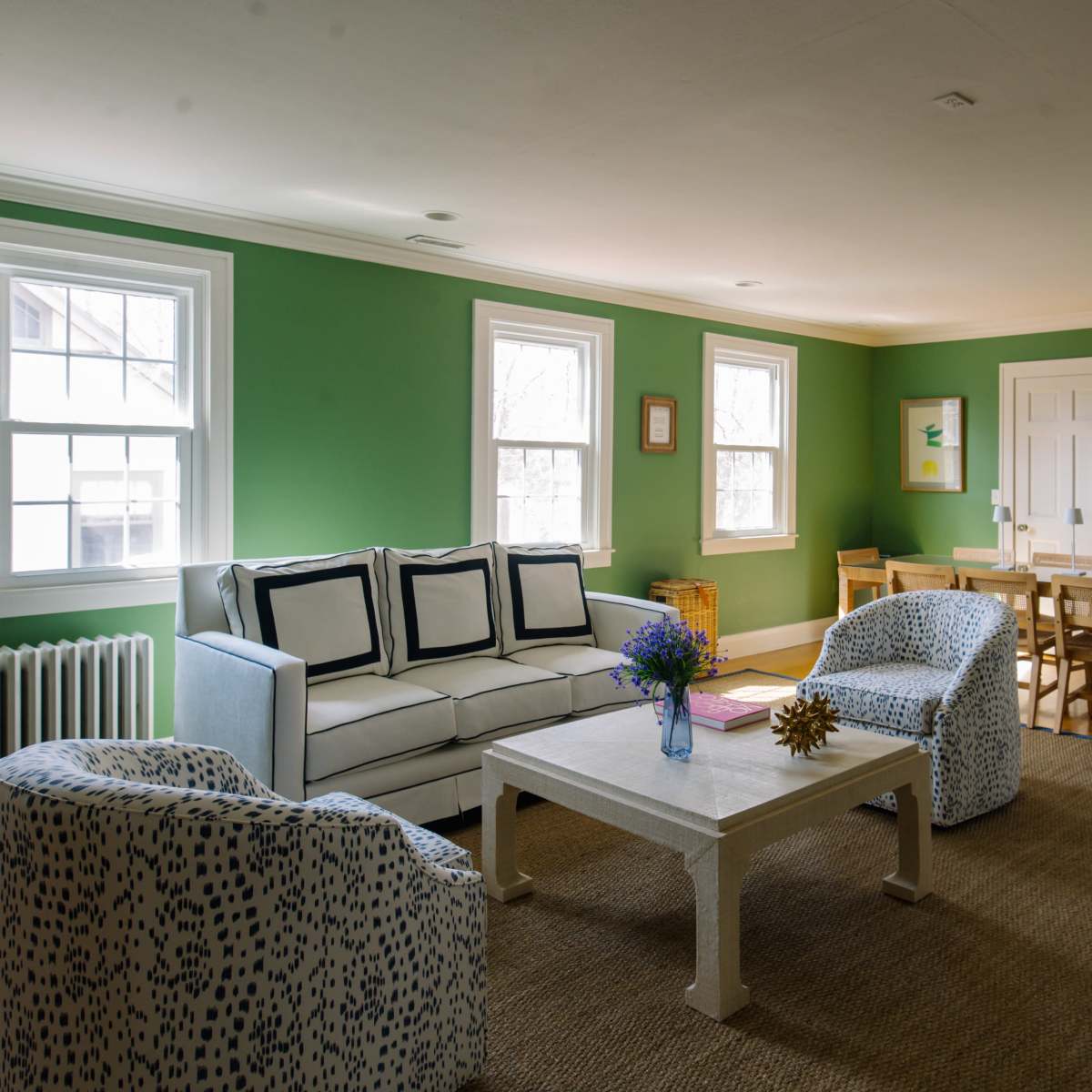 A bright living room features a white couch and two patterned chairs, with a coffee table, surrounded by green walls and ample natural light from multiple windows.