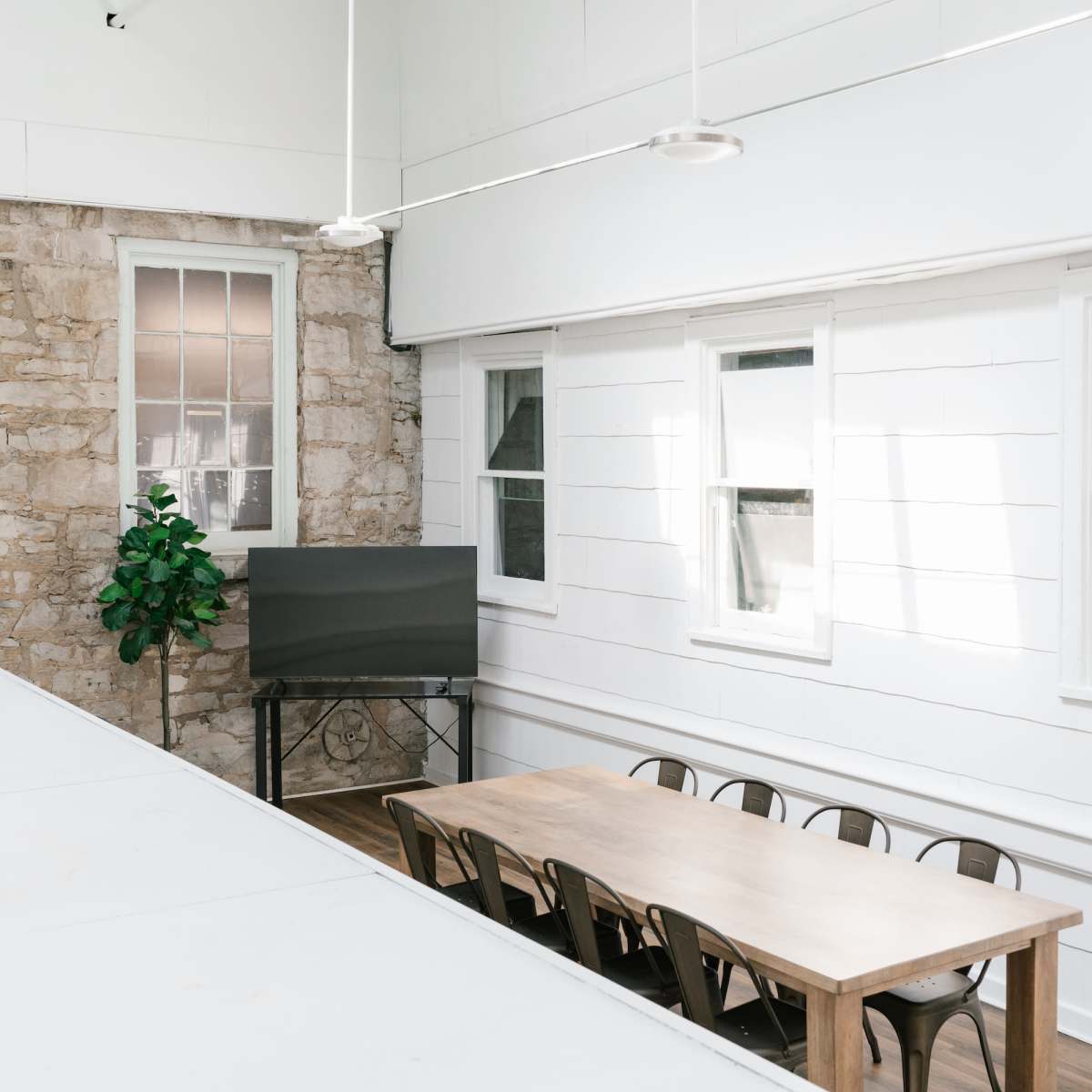 A light-filled room features a long wooden table with metal chairs, a large television, and a potted plant against a stone wall.
