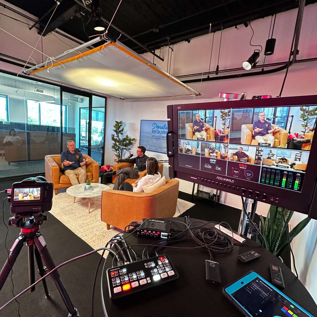 A video production setup features two people seated in chairs while being recorded, with a control monitor displaying multiple camera angles and a mixer visible in the foreground.
