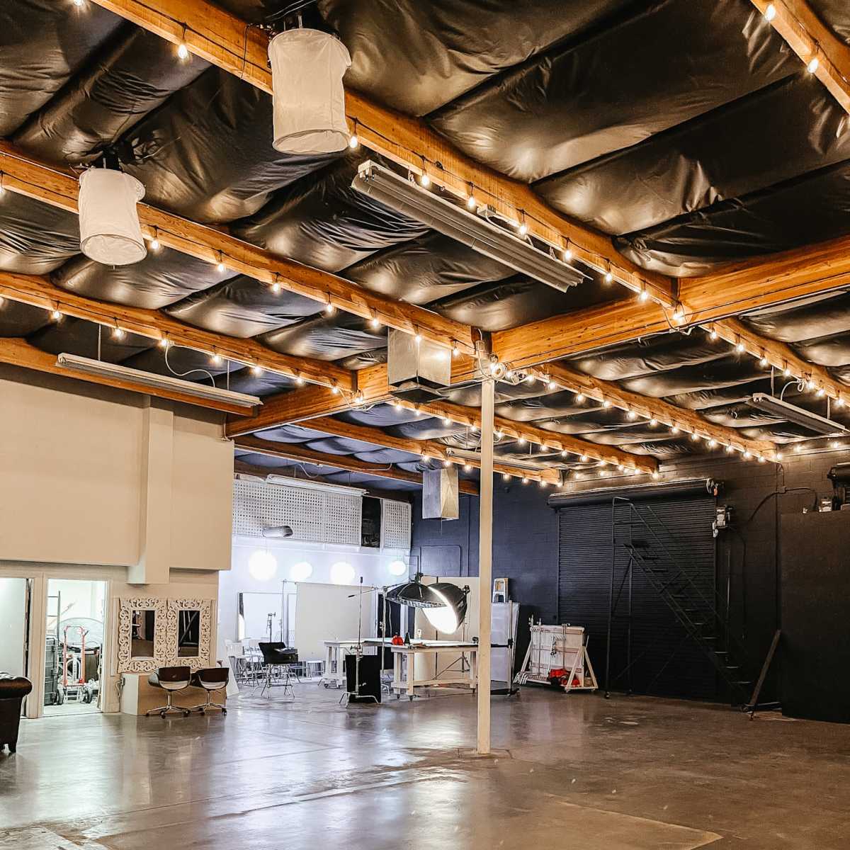 A spacious interior with a polished concrete floor, high ceilings, and string lights overhead, featuring a mix of studio equipment and furniture arranged in a creative workspace.