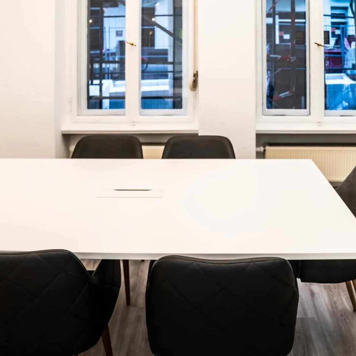 A large white conference table surrounded by six black chairs is positioned in a room with two windows showing an outdoor view.