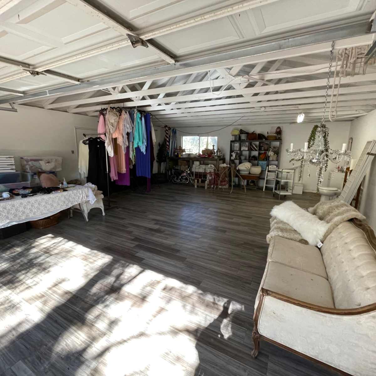 A spacious garage interior with a variety of clothing hanging on racks, a table covered with items, and a vintage couch positioned along one wall.