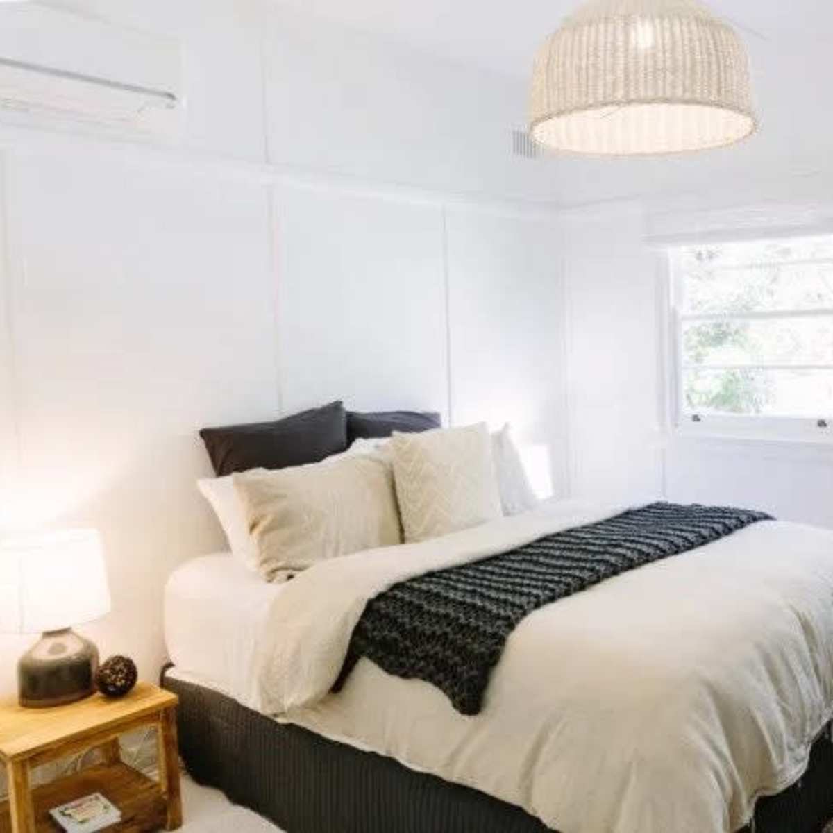 A cozy bedroom features a neatly made bed with cream-colored bedding and a woven throw, accompanied by a bedside lamp and wicker chair against a white-paneled wall.