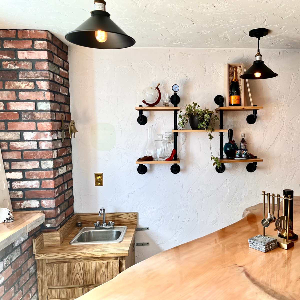 A kitchen area featuring a wooden bar counter, a small sink, and shelves displaying bottles and decorative items against a textured white wall and exposed brick.