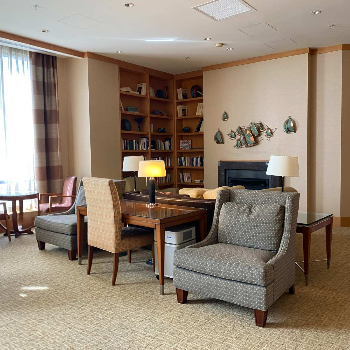 A well-lit lounge area with a bookshelf, comfortable chairs, and a table near a fireplace.