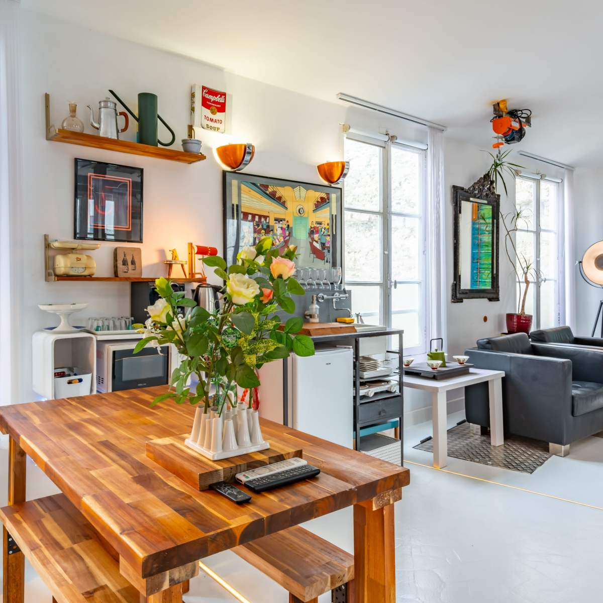 A modern living space features a wooden dining table, black leather sofas, a stylish floor lamp, and decorative art on the walls, surrounded by large windows.