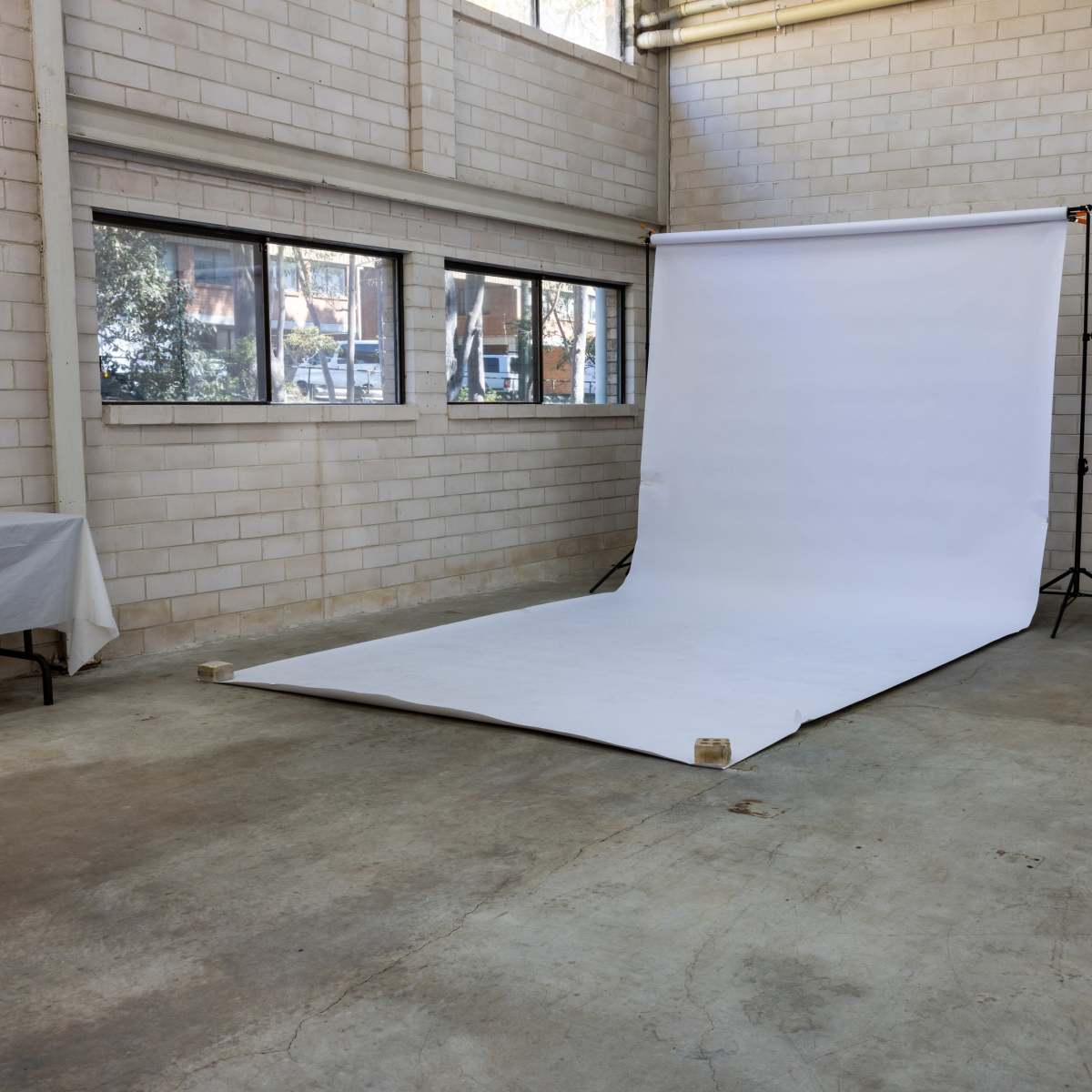 A photography studio features a white backdrop set up on the floor with large windows and bare concrete walls.