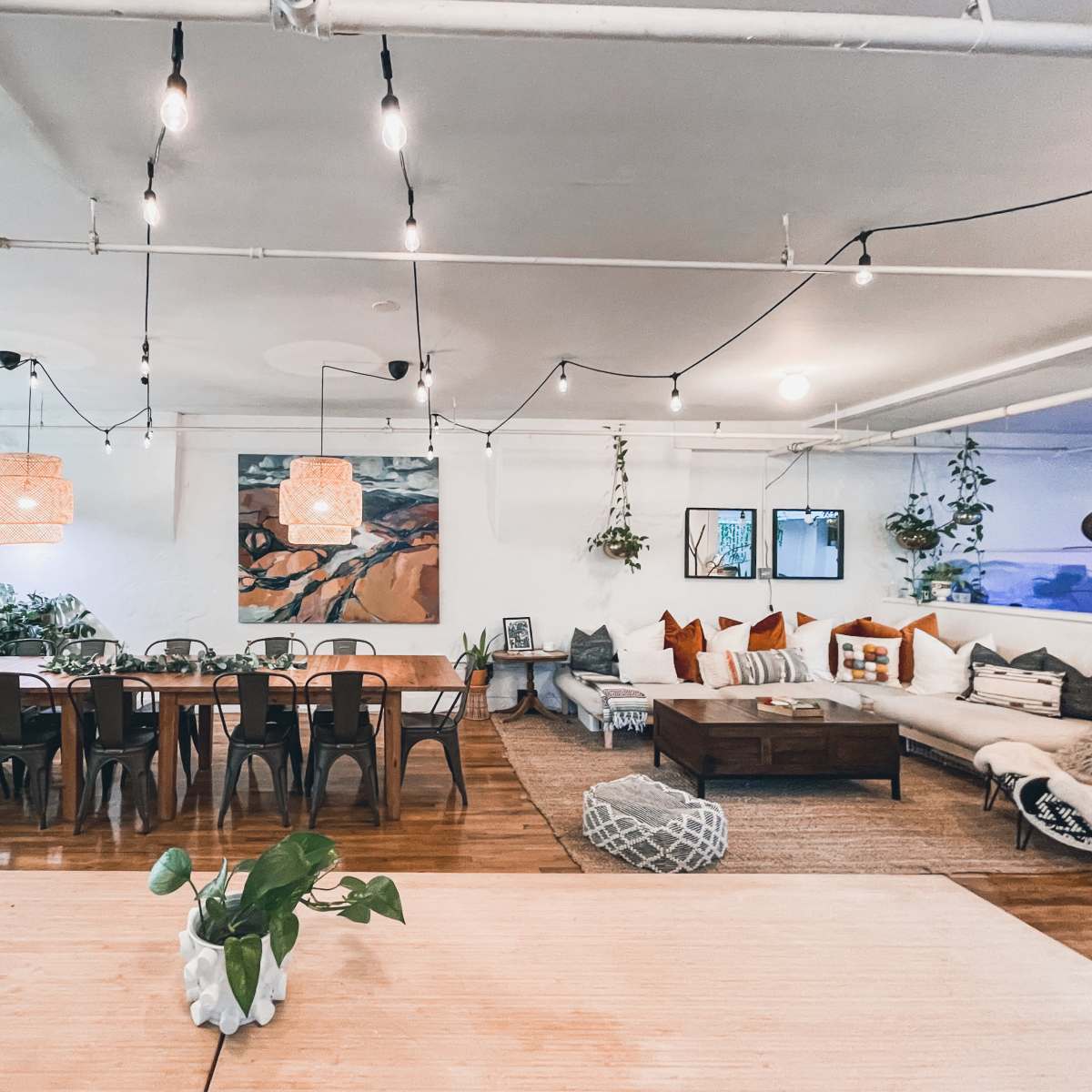 A spacious, light-filled living area with a large dining table, a cozy seating arrangement, decorative plants, and soft lighting fixtures.