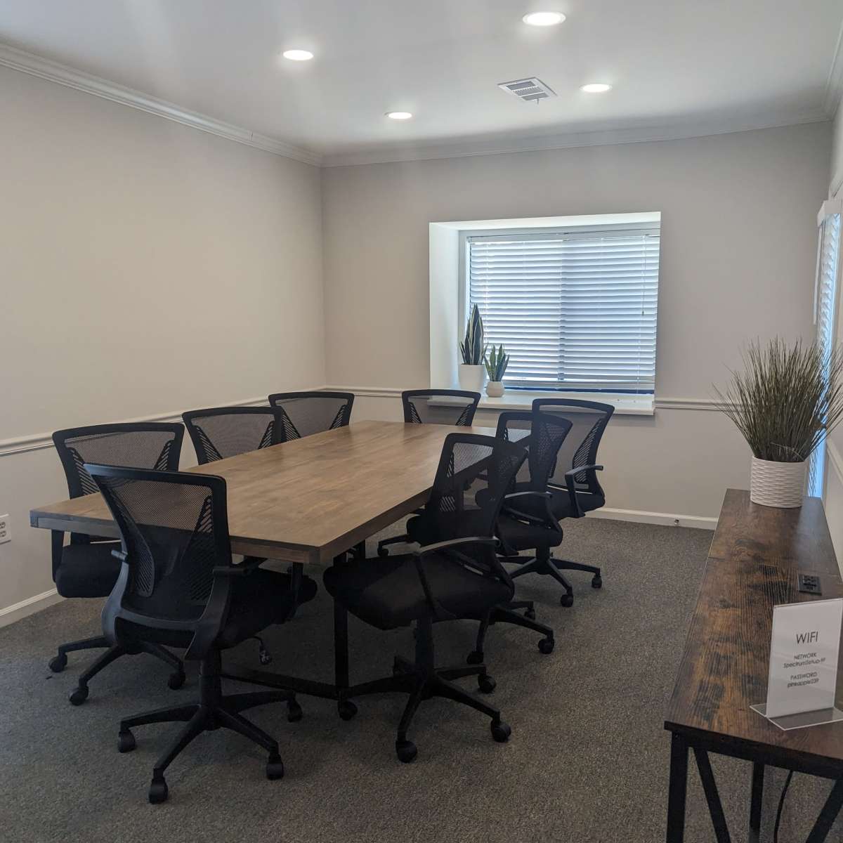 A conference room features a long wooden table surrounded by eight black office chairs, with a window and a plant in the background.