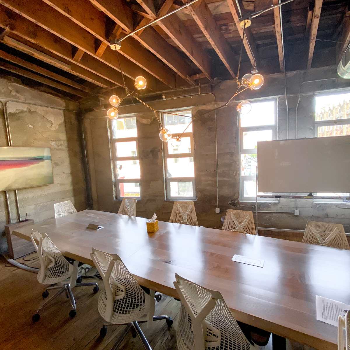 A modern conference room features a long wooden table surrounded by ergonomic chairs, with exposed beams and large windows letting in natural light.