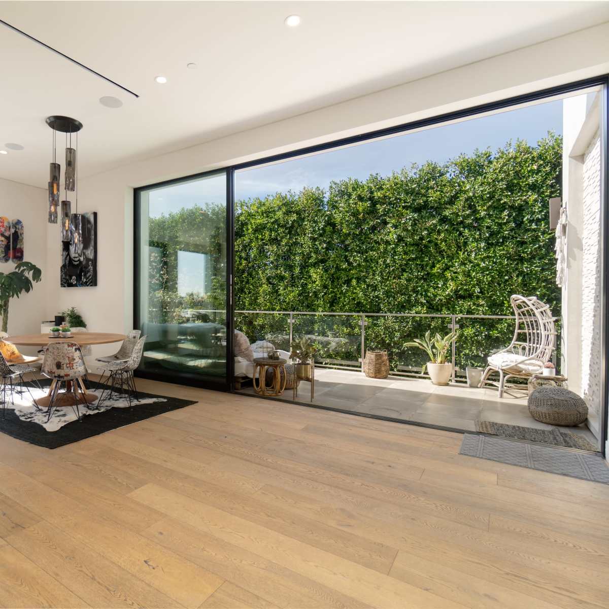 A modern living space with large glass doors that open to a patio surrounded by greenery, featuring a dining area with stylish furniture.