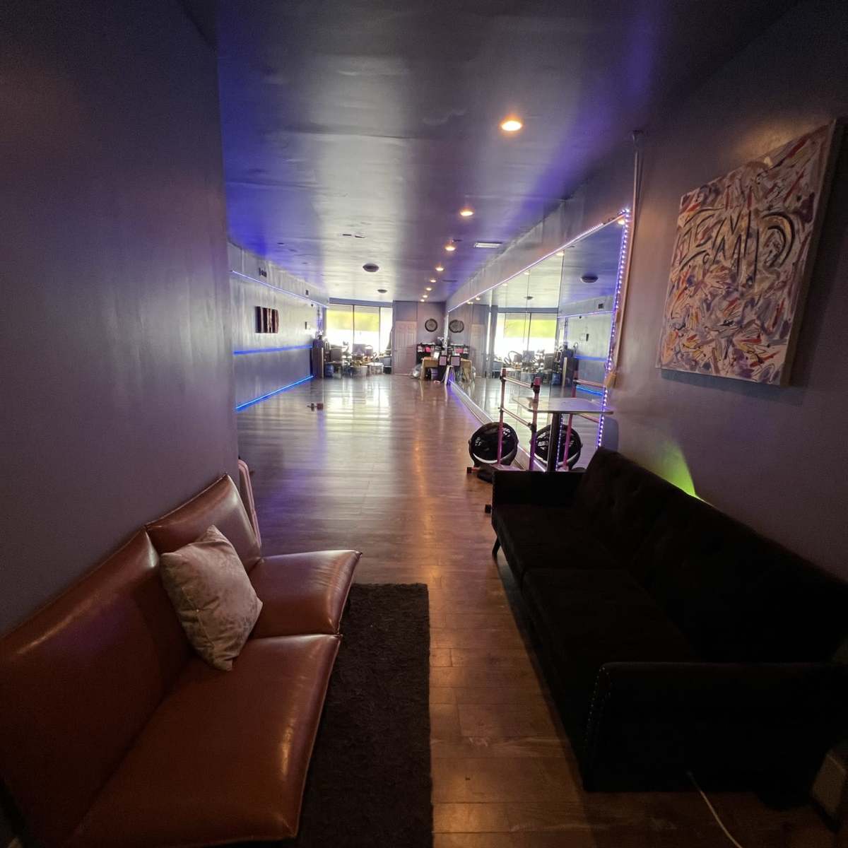 A hallway with two sofas along the side, illuminated by blue lighting, leading towards a spacious area with various furnishings in the background.