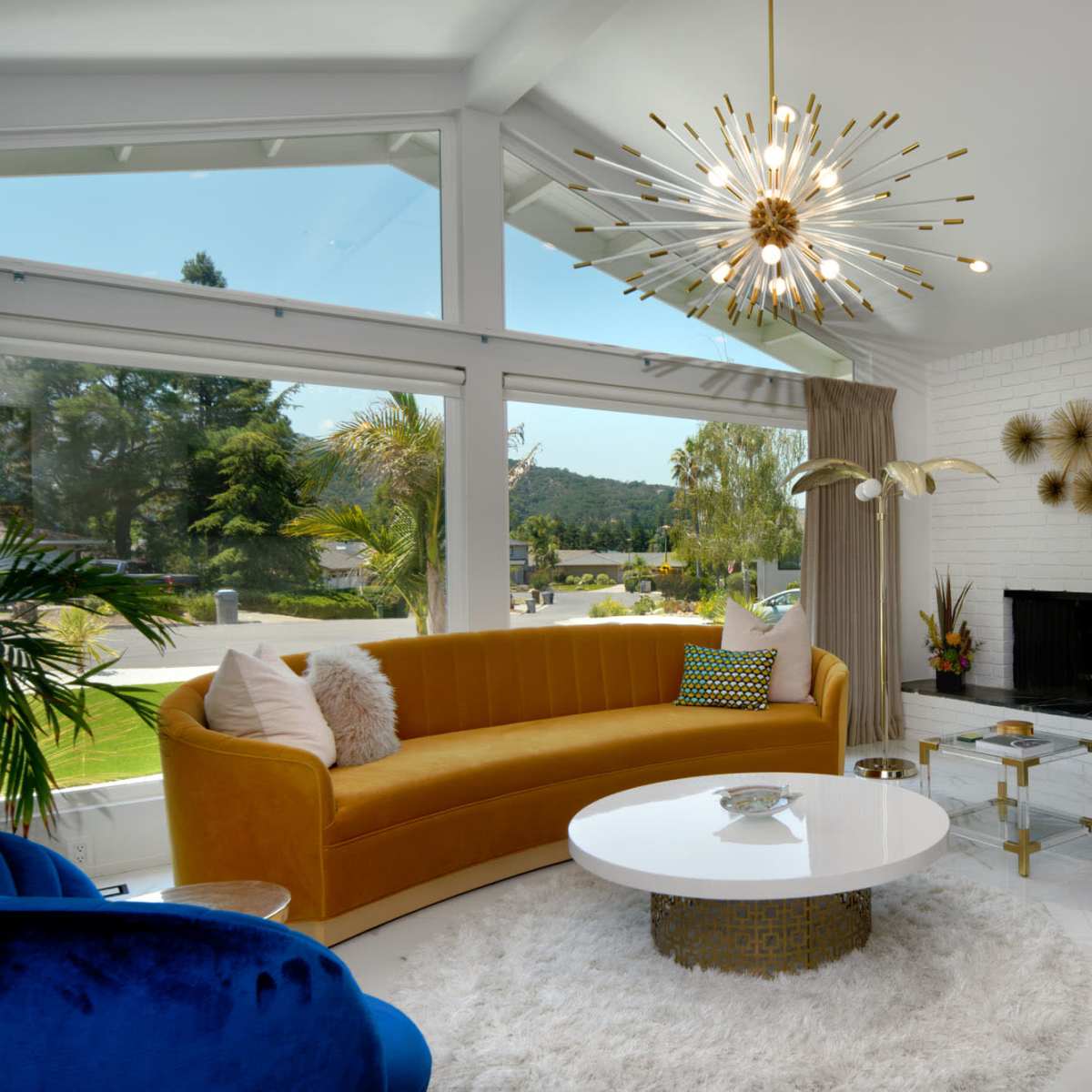A modern living room features a yellow sofa, blue chairs, a round coffee table, and a fireplace, with large windows offering a view of the outdoors.