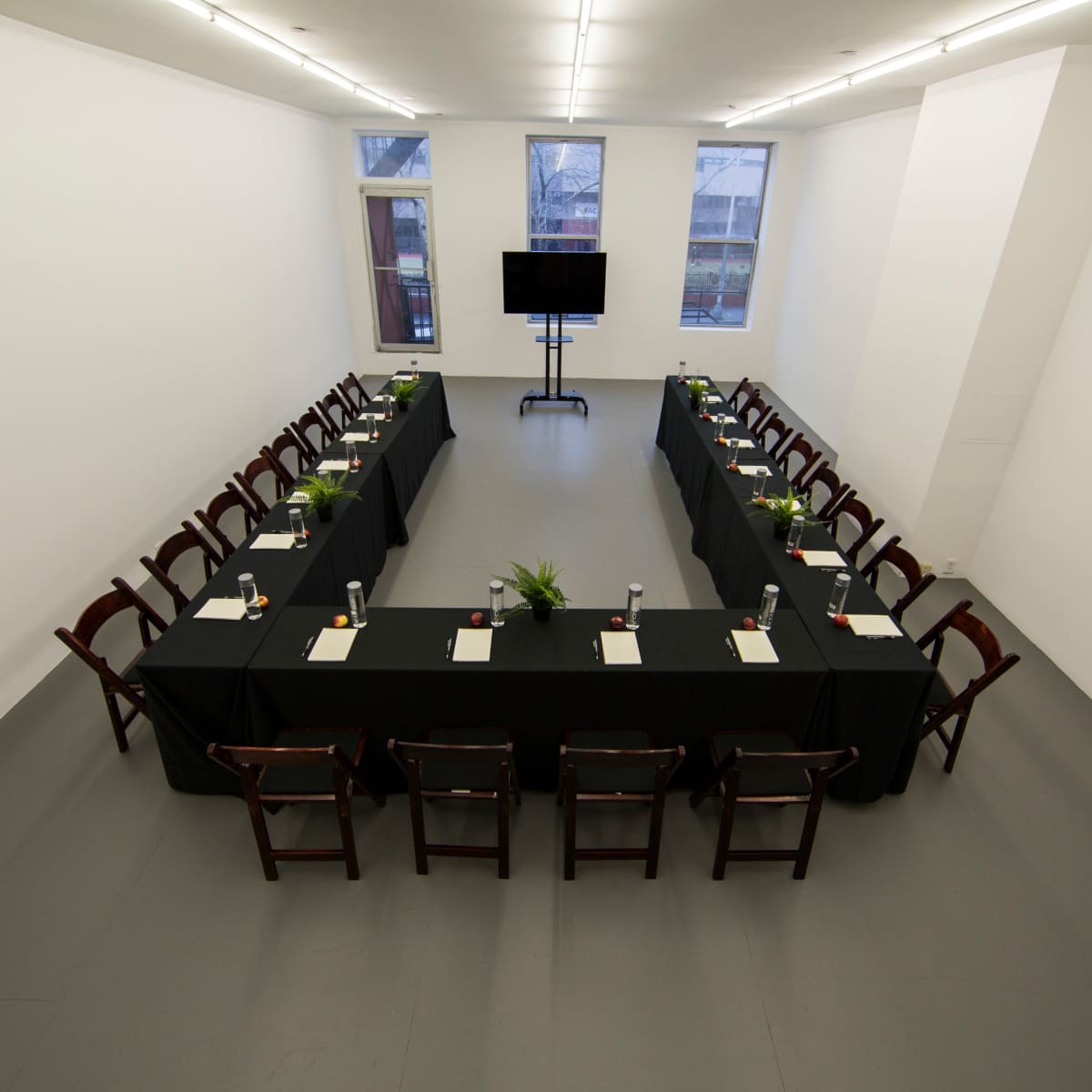 A rectangular conference room features a long table set with tableware and surrounded by wooden chairs, with a television screen at one end and large windows on the walls.