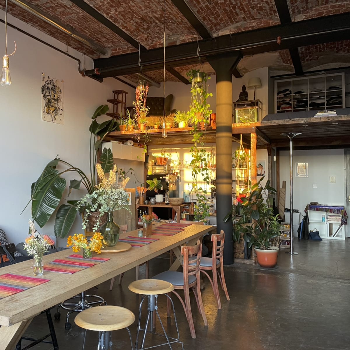 A spacious, industrial-style dining area with a long wooden table, surrounded by plants and decorative elements, along with an open kitchen area in the background.