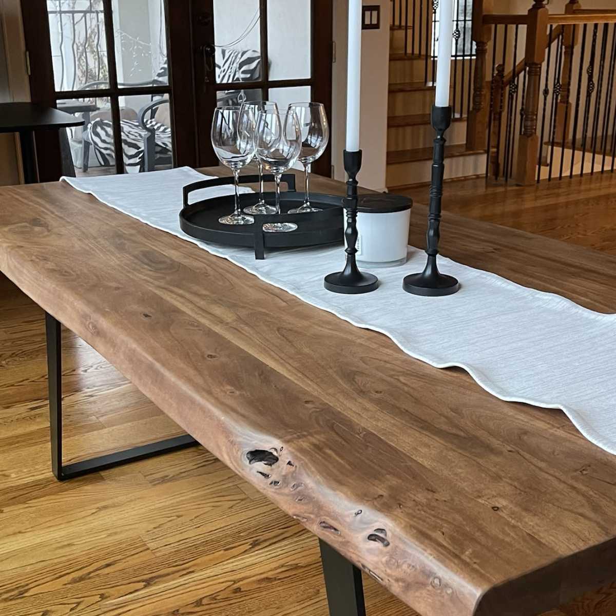 A wooden dining table with a natural edge is set with a white runner, two candles, a bowl, and a tray of glasses.