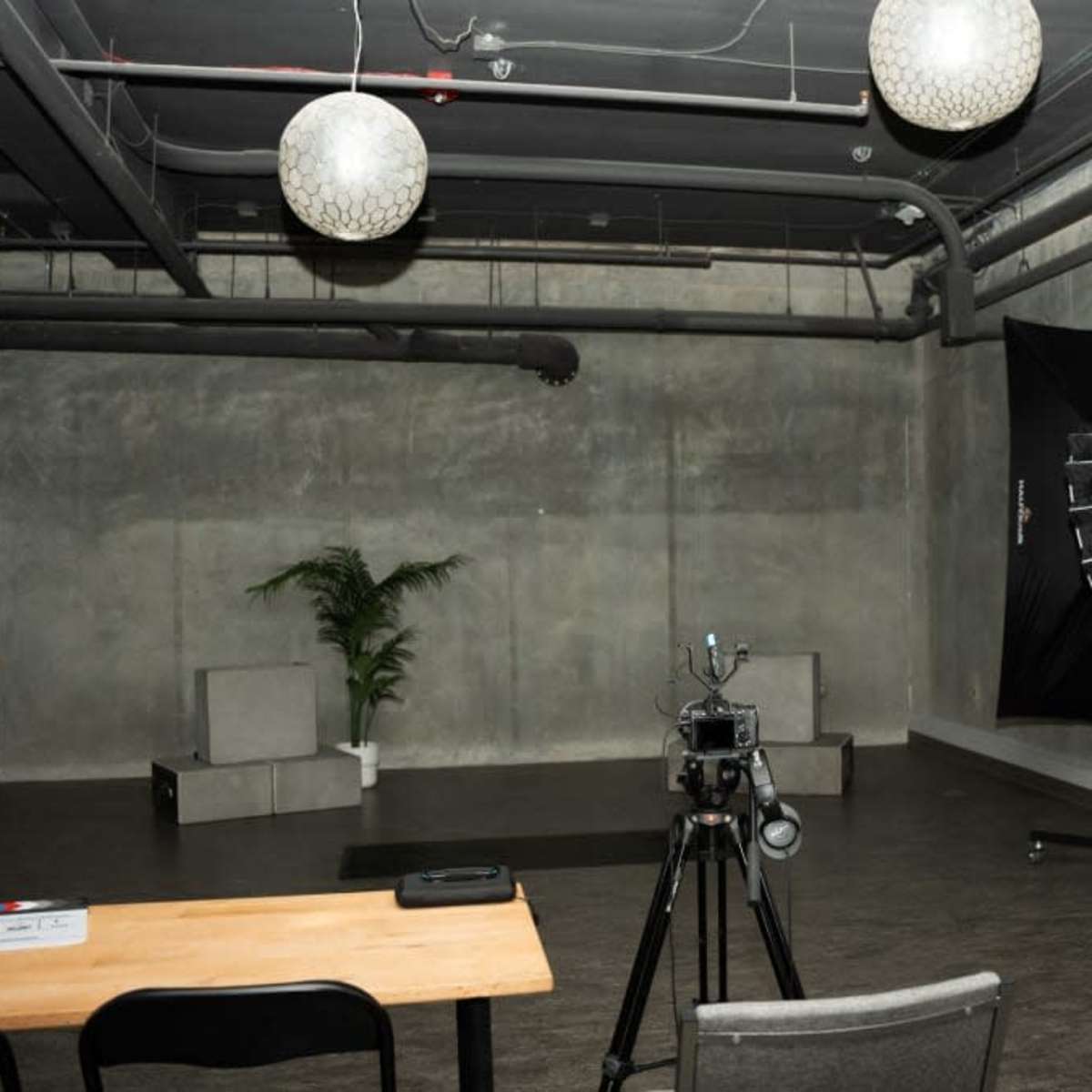 A minimalist studio space with a camera on a tripod, softbox lighting, a whiteboard, and a potted plant in a concrete-finished room.