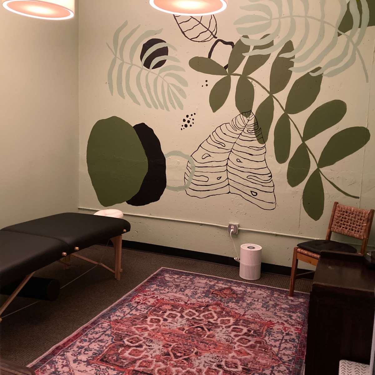 A small room with a massage table, a patterned rug, and a mural of plants on the wall.