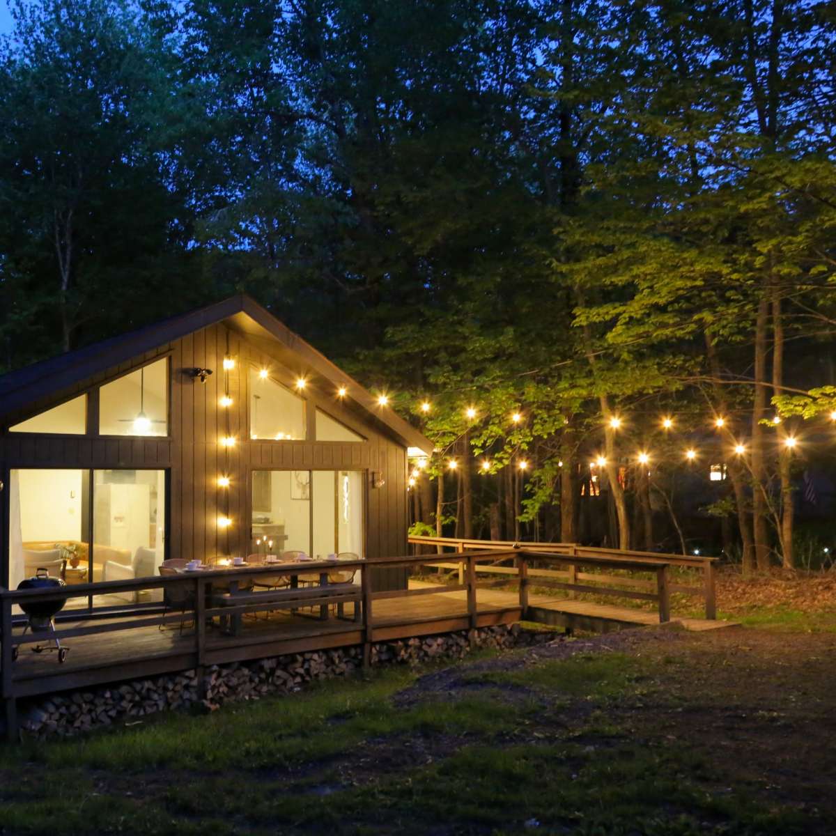 A modern wooden cabin with large windows is illuminated by string lights outside, situated in a wooded area during twilight.