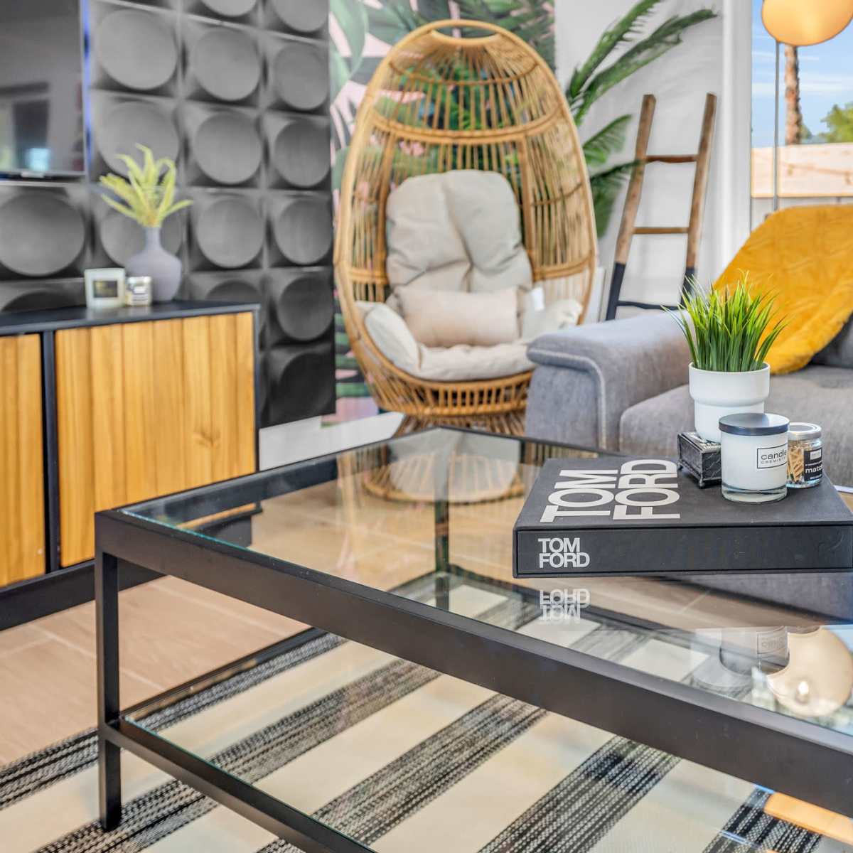 The scene features a modern living room with a glass coffee table topped with books and a decorative plant, surrounded by a gray sofa and a wicker chair.