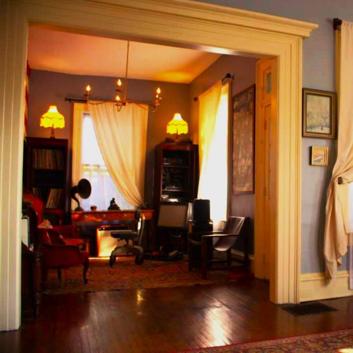 A cozy interior with two adjoining rooms, featuring large windows draped with white curtains and vintage furniture, including a console table and seating arrangements.