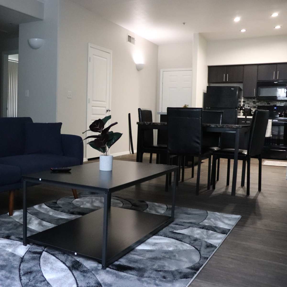 A modern living room with a blue sofa, a black coffee table on a patterned rug, and a dining area in the background.