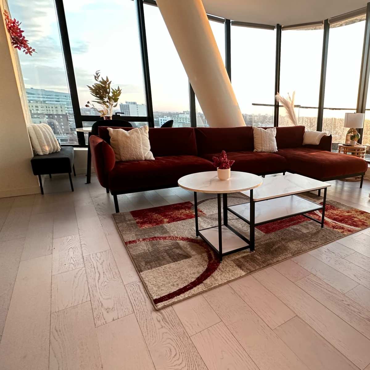A modern living room with a red sofa, a small coffee table on a patterned rug, and a decorated Christmas tree by large windows that overlook a sunset.