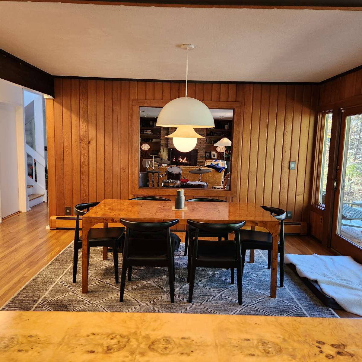 A dining room features a wooden table surrounded by black chairs, with a large pendant light above and wooden wall panels.