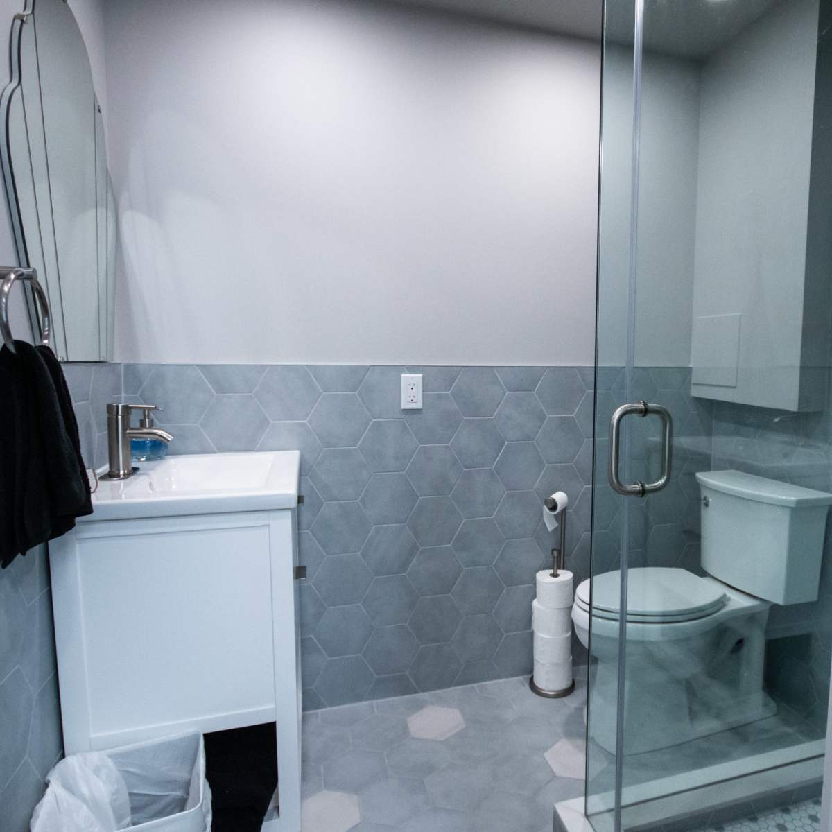 A modern bathroom featuring a glass shower, a white toilet, a sink with a mirror, and hexagonal tiled flooring.