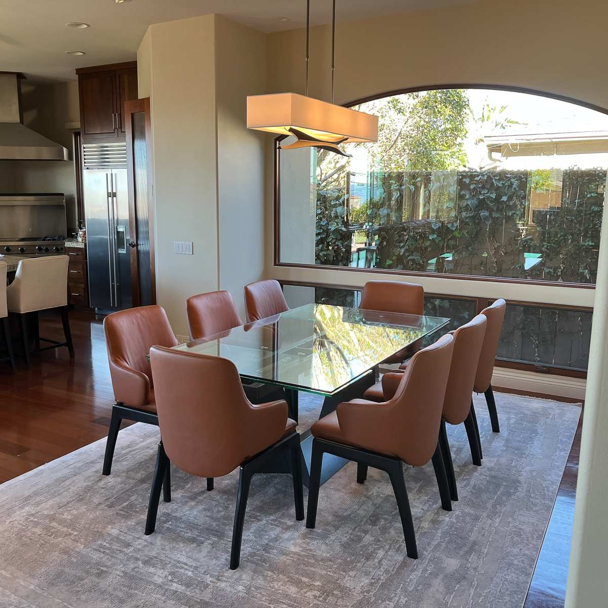 A glass dining table surrounded by brown chairs is positioned on a rug in a well-lit dining area that opens to a garden view.