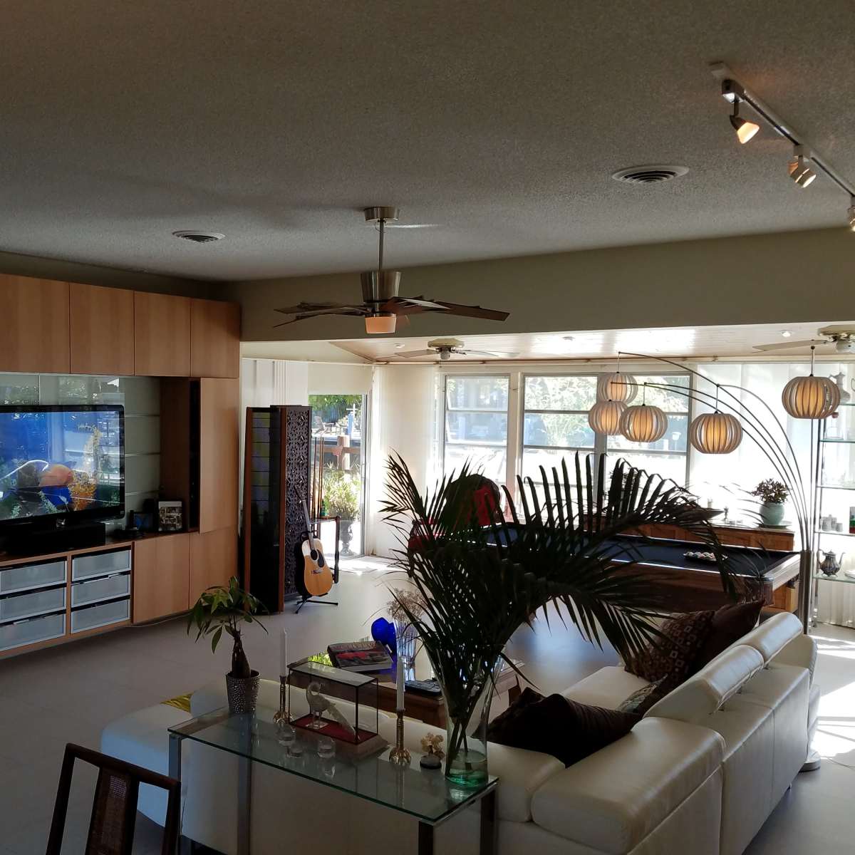 The scene shows a modern living room with a large sectional sofa, a wall-mounted television, and large windows allowing natural light to enter the space.