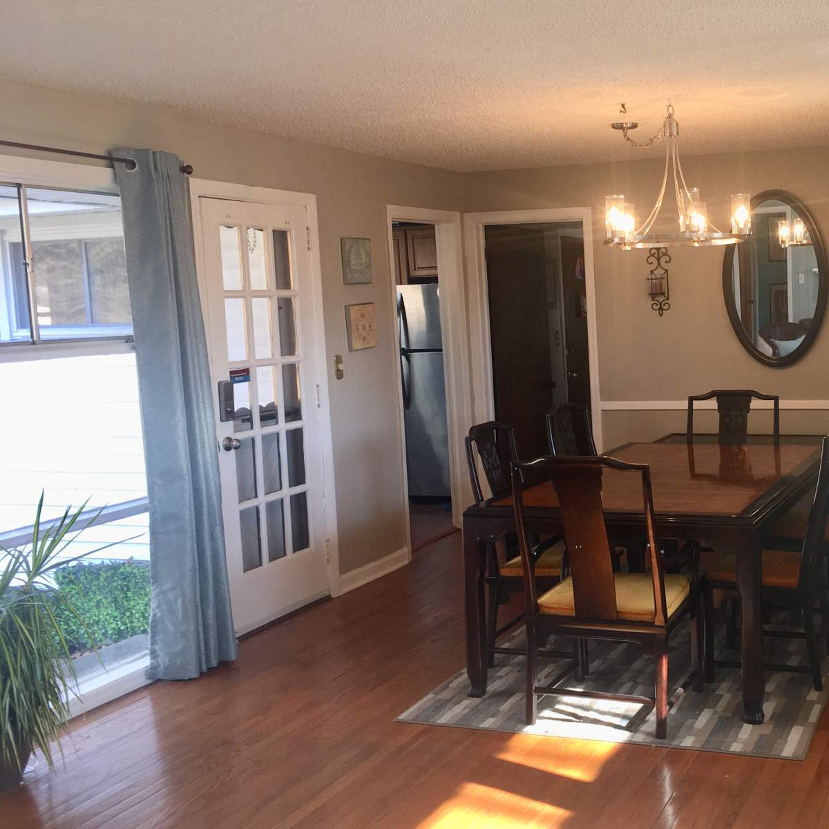 A dining area with a wooden table surrounded by chairs, a chandelier above, and a mirror on the wall, adjacent to a kitchen area and a door leading outside.