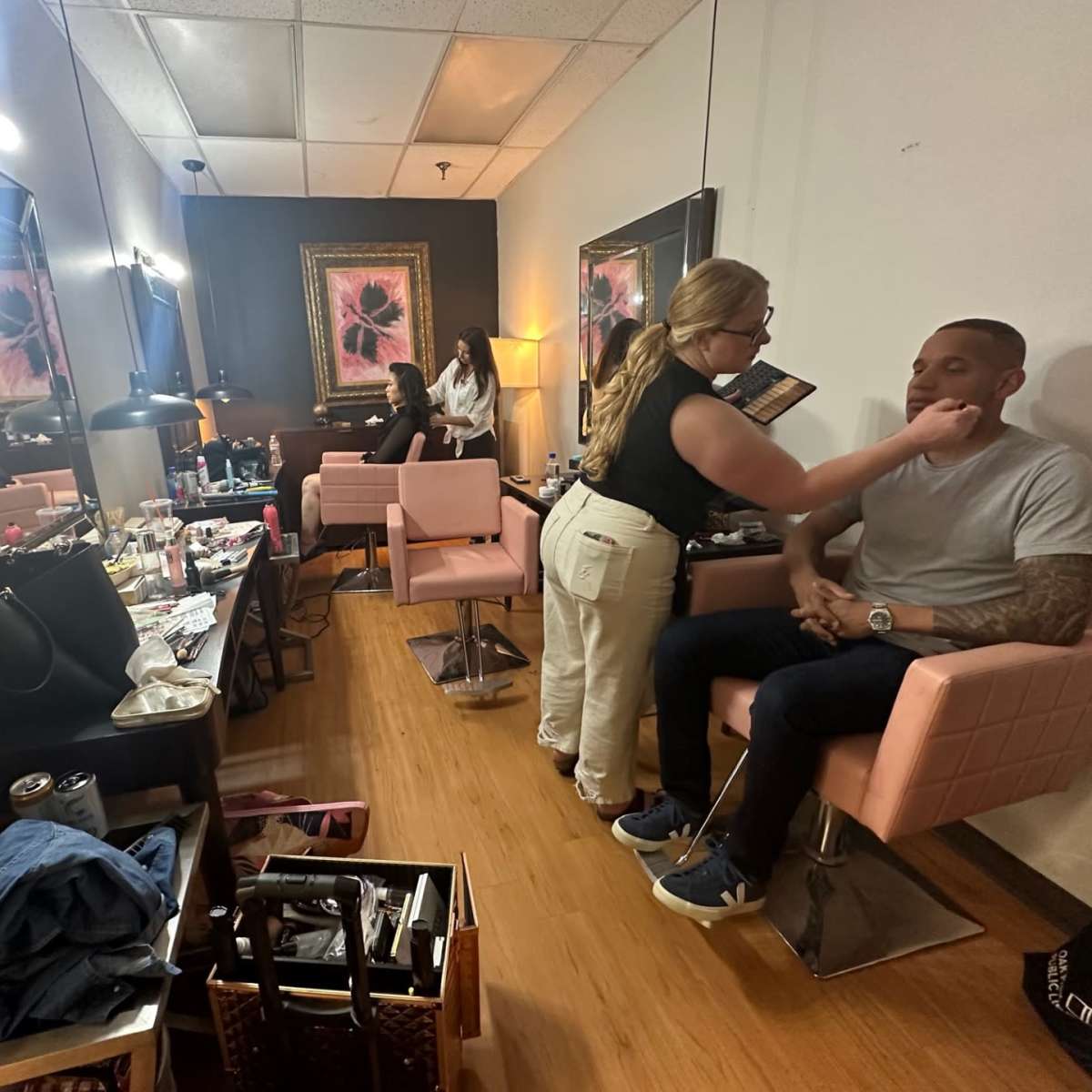 A makeup artist applies makeup to a seated man in a well-lit dressing room featuring mirrors and various beauty products.